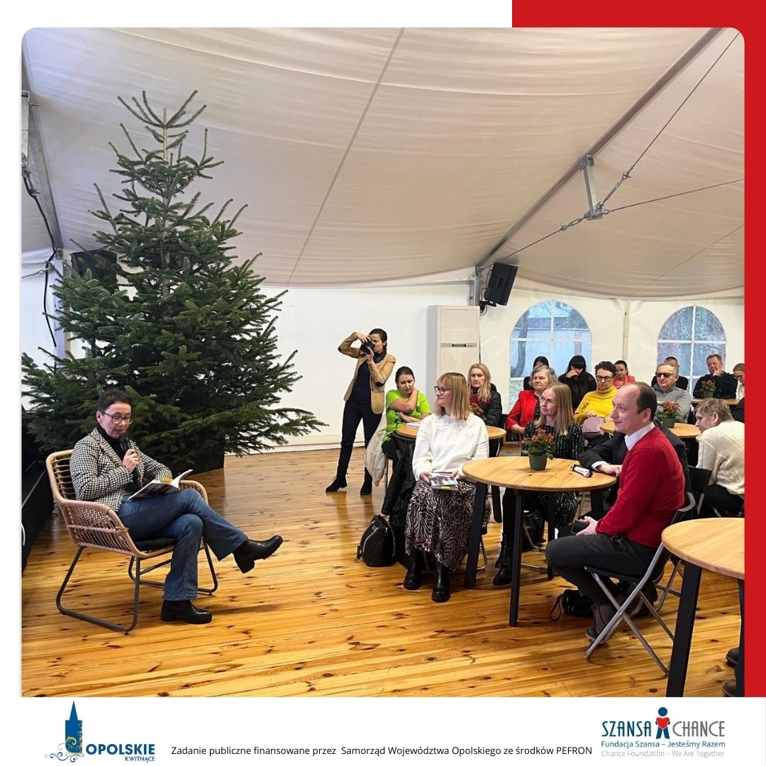 Kobieta w jeansach siedzi na wiklinowym krześle i czyta fragment książki. W tle widoczna jest choinka, a w pierwszym planie uczestnicy siedzący przy okrągłych stolikach, z których niektóre są udekorowane doniczkami z czerwonymi kwiatami. W dolnej części zdjęcia znajdują się logotypy: Opolskie Kwitnące oraz Fundacja Szansa- Jesteśmy Razem. Tekst informuje, że zadanie jest finansowane przez Samorząd Województwa Opolskiego ze środków PFRON.