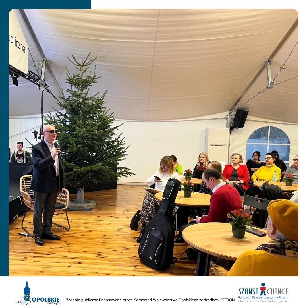 Mężczyzna w ciemnym garniturze i okularach przeciwsłonecznych przemawia na scenie w namiocie. W tle widoczna jest choinka, a w pierwszym planie uczestnicy siedzący przy okrągłych stolikach, z których niektóre są udekorowane doniczkami z czerwonymi kwiatami. W dolnej części zdjęcia znajdują się logotypy: Opolskie Kwitnące oraz Fundacja Szansa- Jesteśmy Razem. Tekst informuje, że zadanie jest finansowane przez Samorząd Województwa Opolskiego ze środków PFRON.